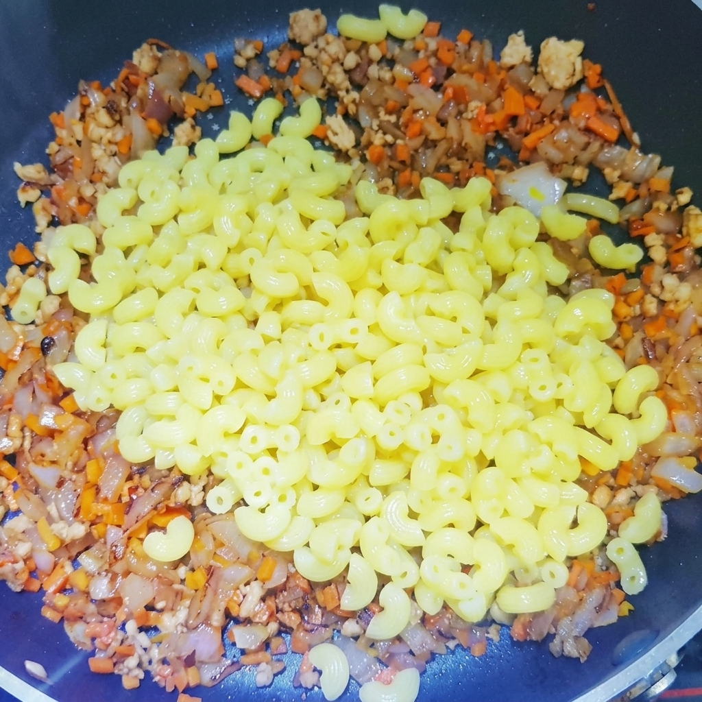 Boiled macaroni being added to a pan filled with a cooked base of minced meat, onions, and carrots.