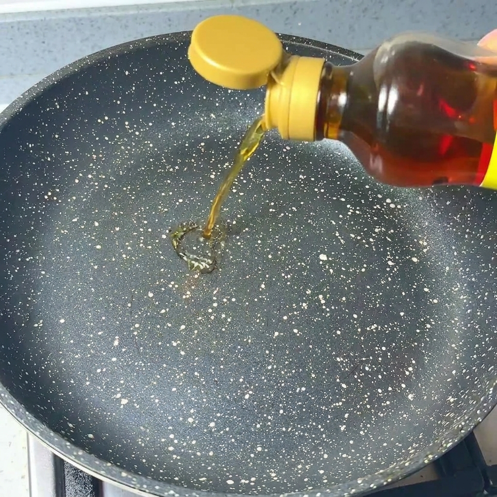 Clear sesame oil being poured from a bottle into a dark, speckled frying pan.