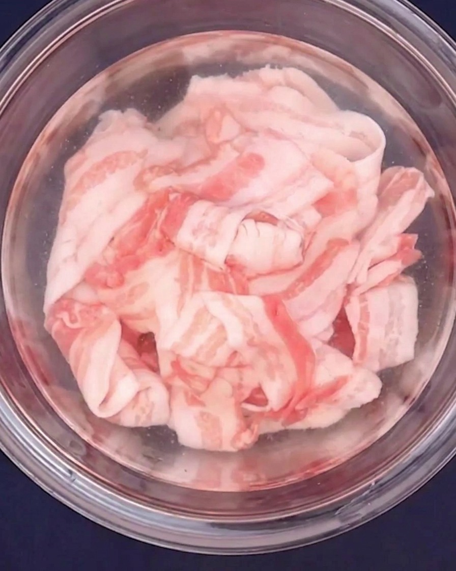 Thinly sliced pork belly being blanched with hot water in a glass bowl.
