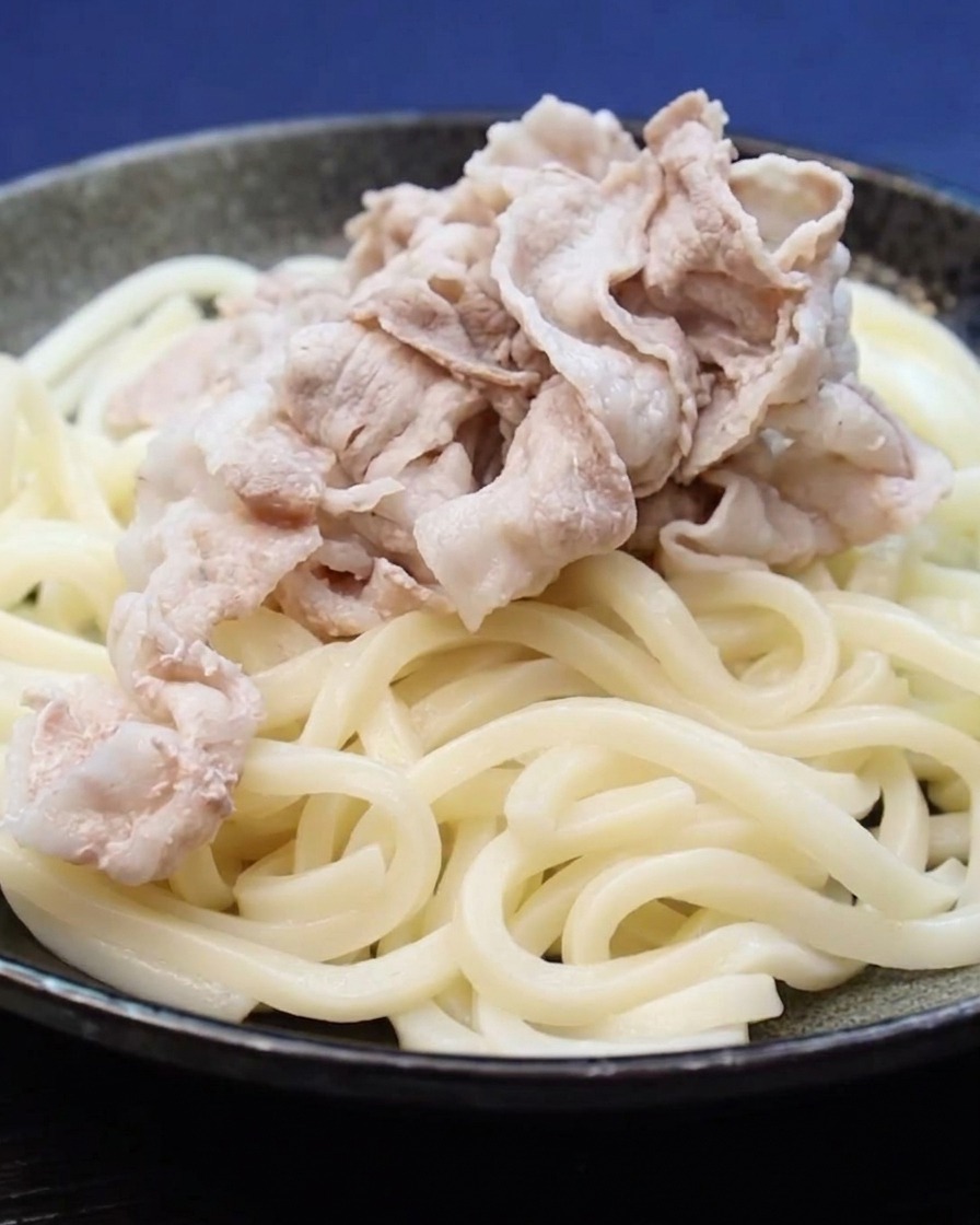 Cooked pork belly slices being placed on a bed of udon noodles.