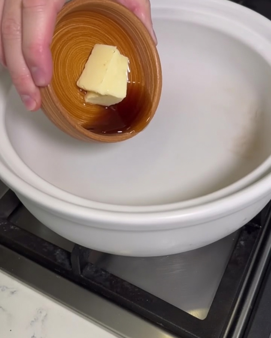 A hand adding a block of butter and liquid sesame oil from a small wooden bowl into an empty white clay pot.