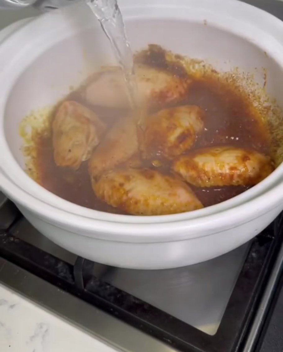 Clear water being poured from a vessel into a bubbling clay pot filled with curry chicken wings.