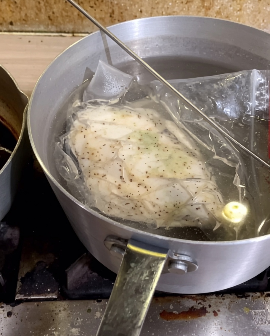 A vacuum-sealed bag containing seasoned chicken breast submerged in a pot of hot water.