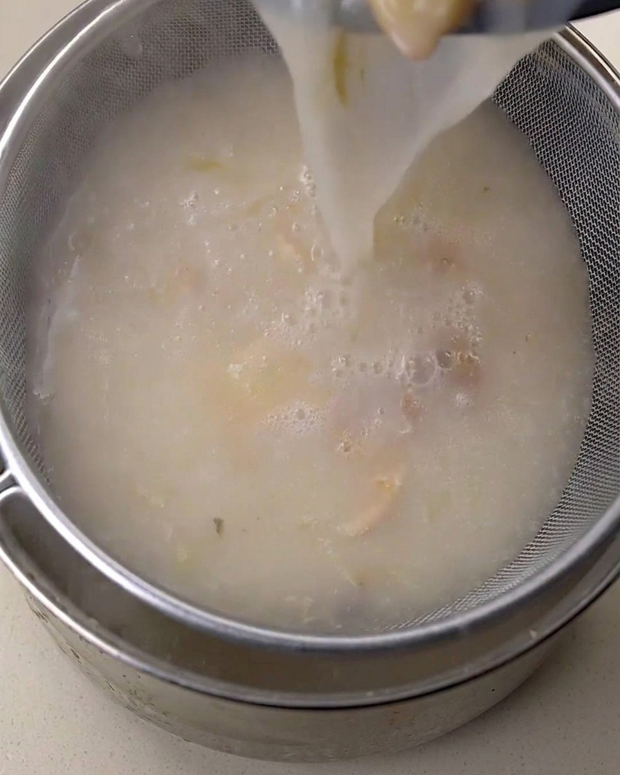 Creamy white chicken broth being poured through a fine-mesh sieve into a metal container.