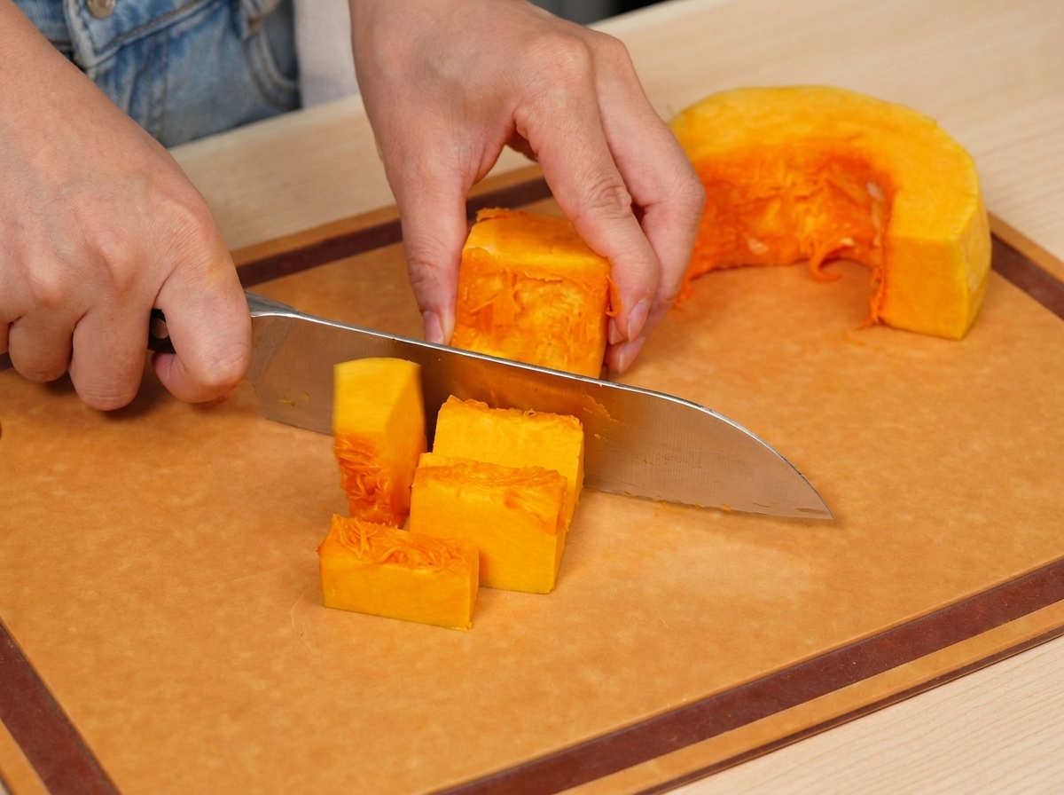 Fresh orange pumpkin being carefully sliced into smaller, even cubes on a wooden cutting board.