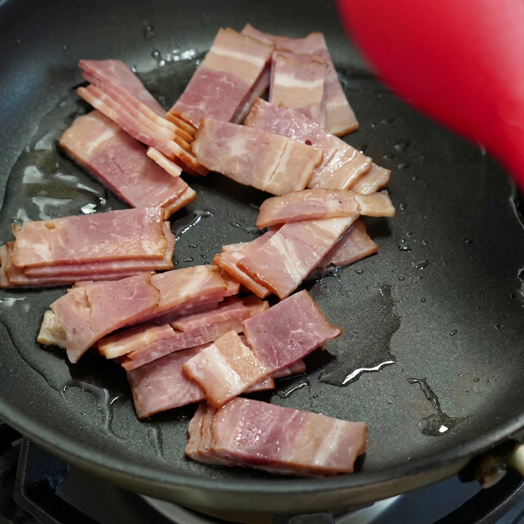 Bacon slices cooking and browning in a black skillet, being stirred with a red spatula.