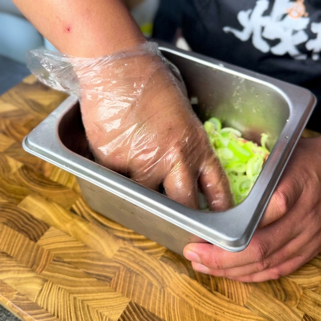 A gloved hand mixing chopped scallions and marinated beef inside a metal container.