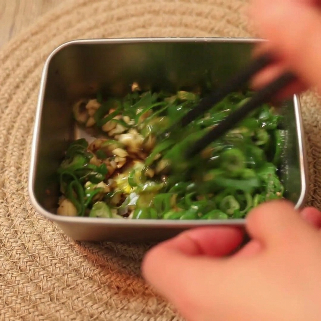 Black chopsticks stirring a mixture of green scallions, garlic, and soy sauce in a metal container.