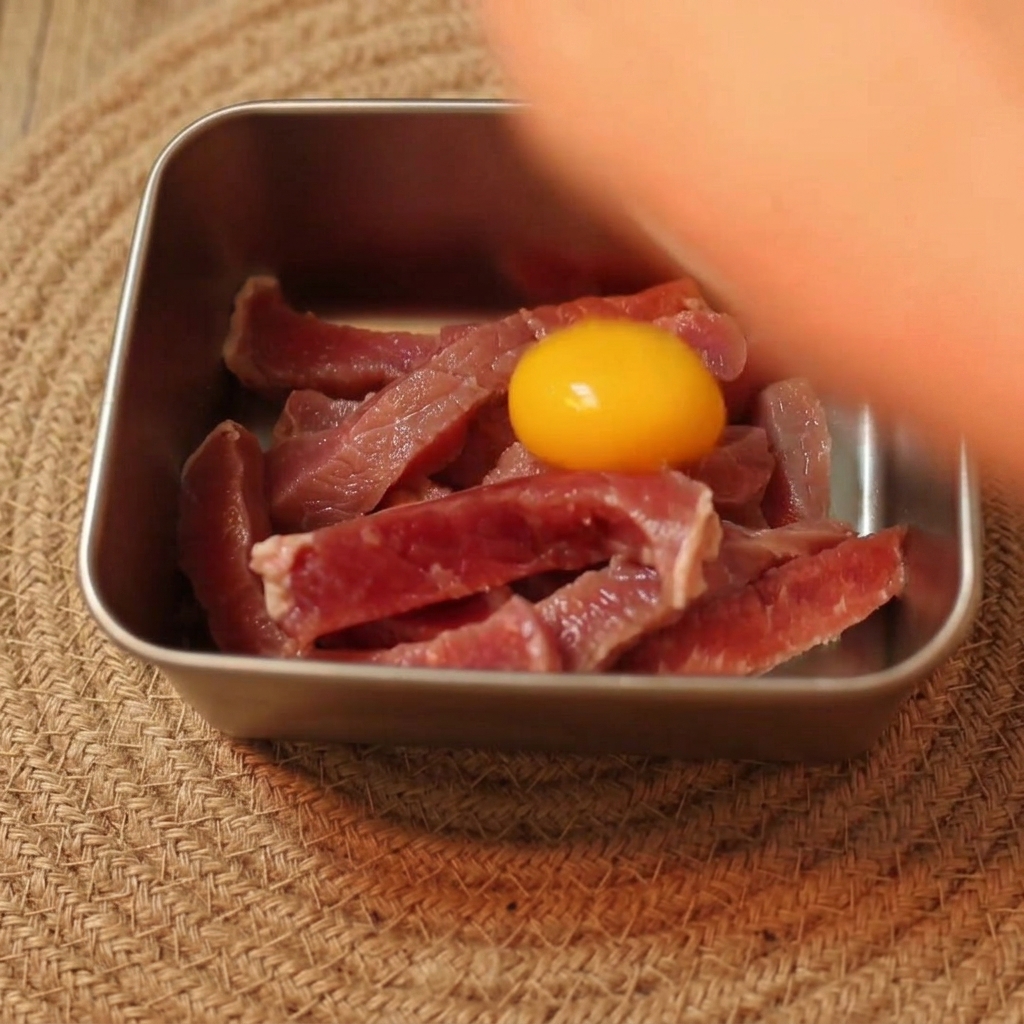 A bright yellow raw egg yolk resting on top of sliced raw beef strips in a metal prep bowl.