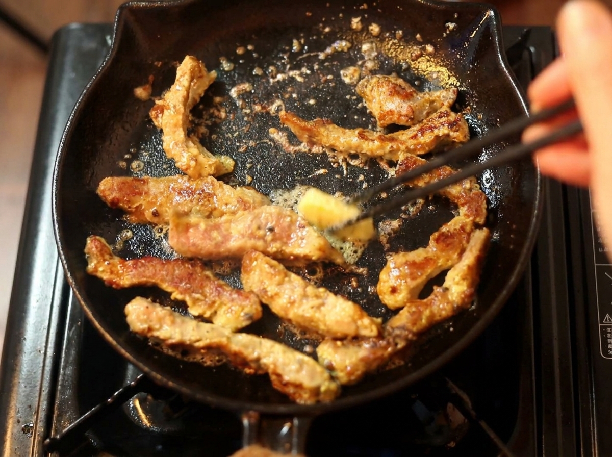 A small piece of butter being added with chopsticks to partially cooked beef frying in a black cast iron skillet.