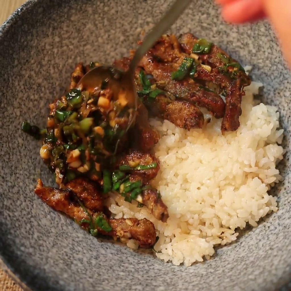 A spoon serving hot cooked beef and green scallion sauce over a bed of white rice in a ceramic bowl.