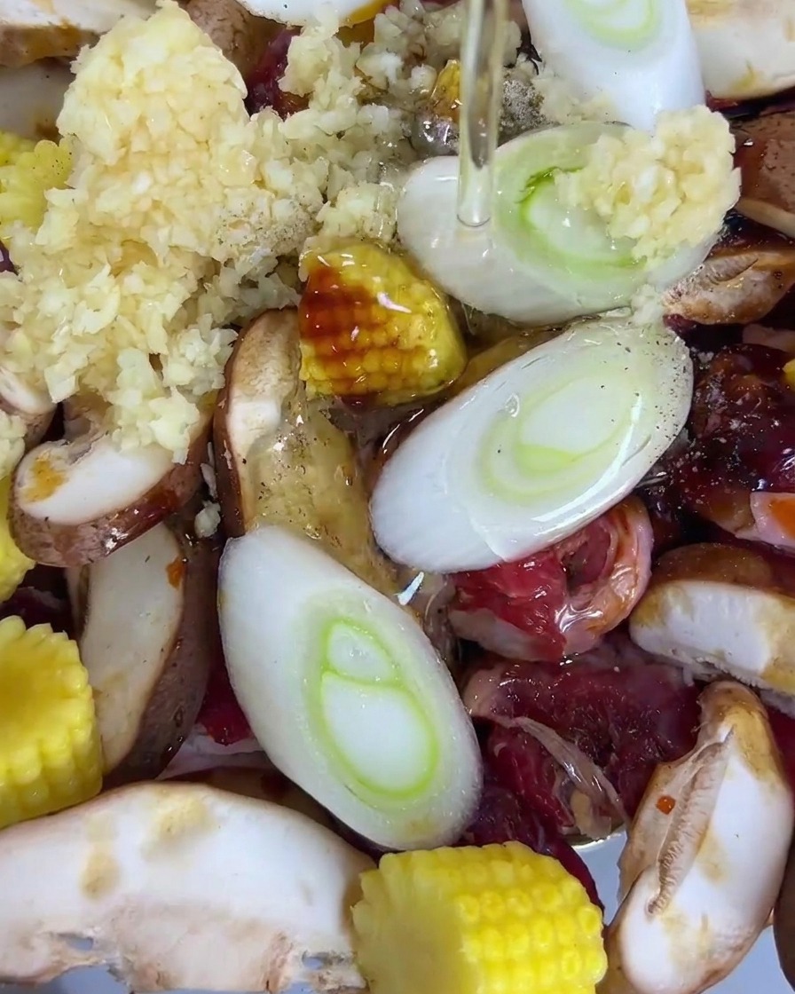 Cooking oil being poured over a tray containing sliced beef, shiitake mushrooms, baby corn, green onions, and garlic.