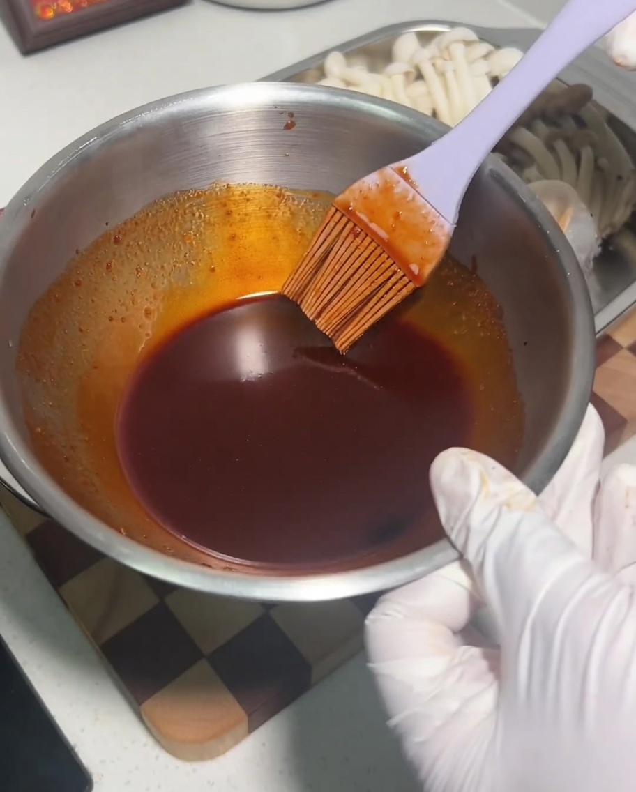 A metal bowl containing a dark, spicy sauce mixture being stirred with a silicone brush.