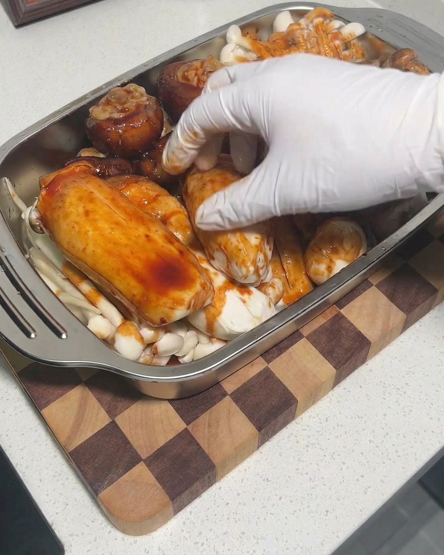 Gloved hands massaging sauce onto the mushrooms in a tray to ensure thorough coating.