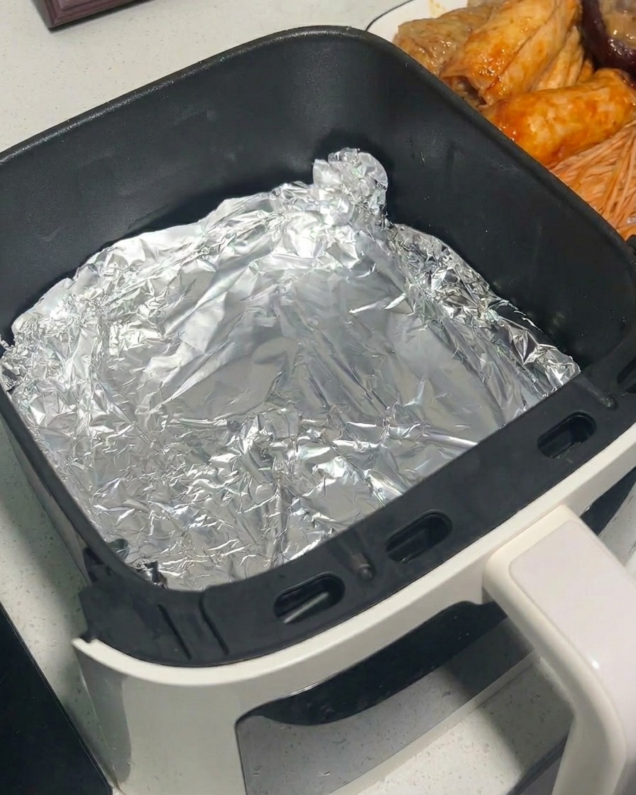 Marinated mushrooms being placed into an air fryer basket lined with aluminum foil using metal tongs.