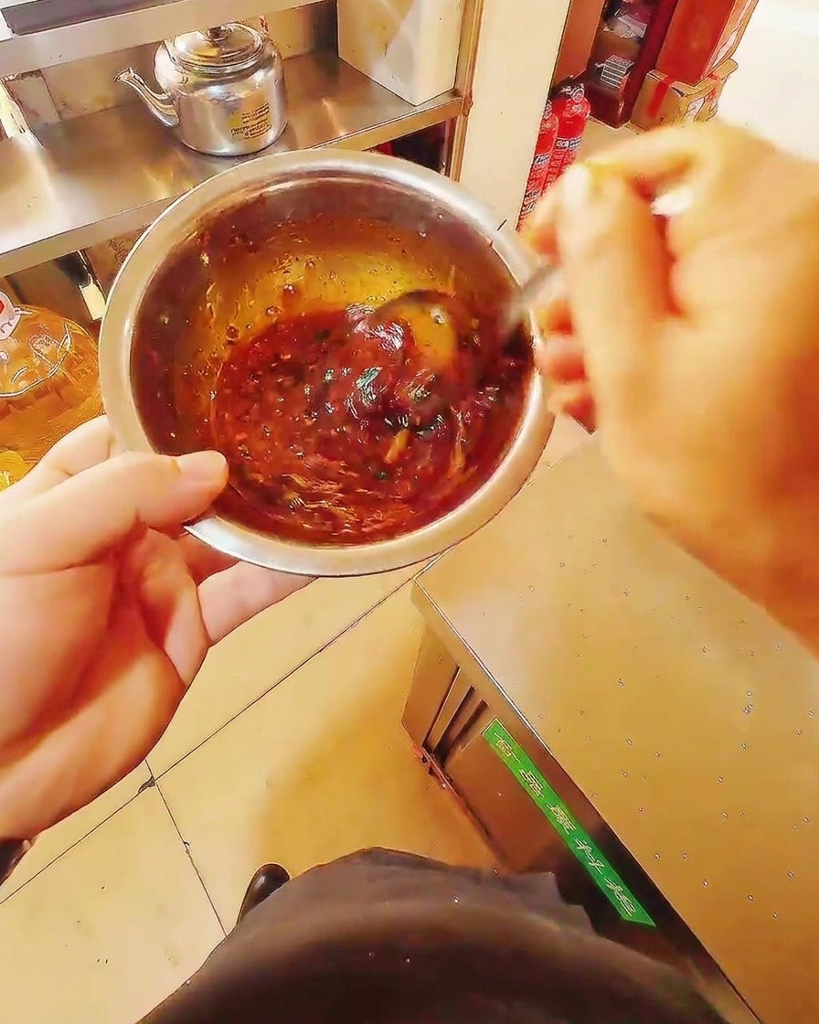 A close-up of a stainless steel bowl containing a rich, dark-red marinade mixture being stirred with a metal spoon.