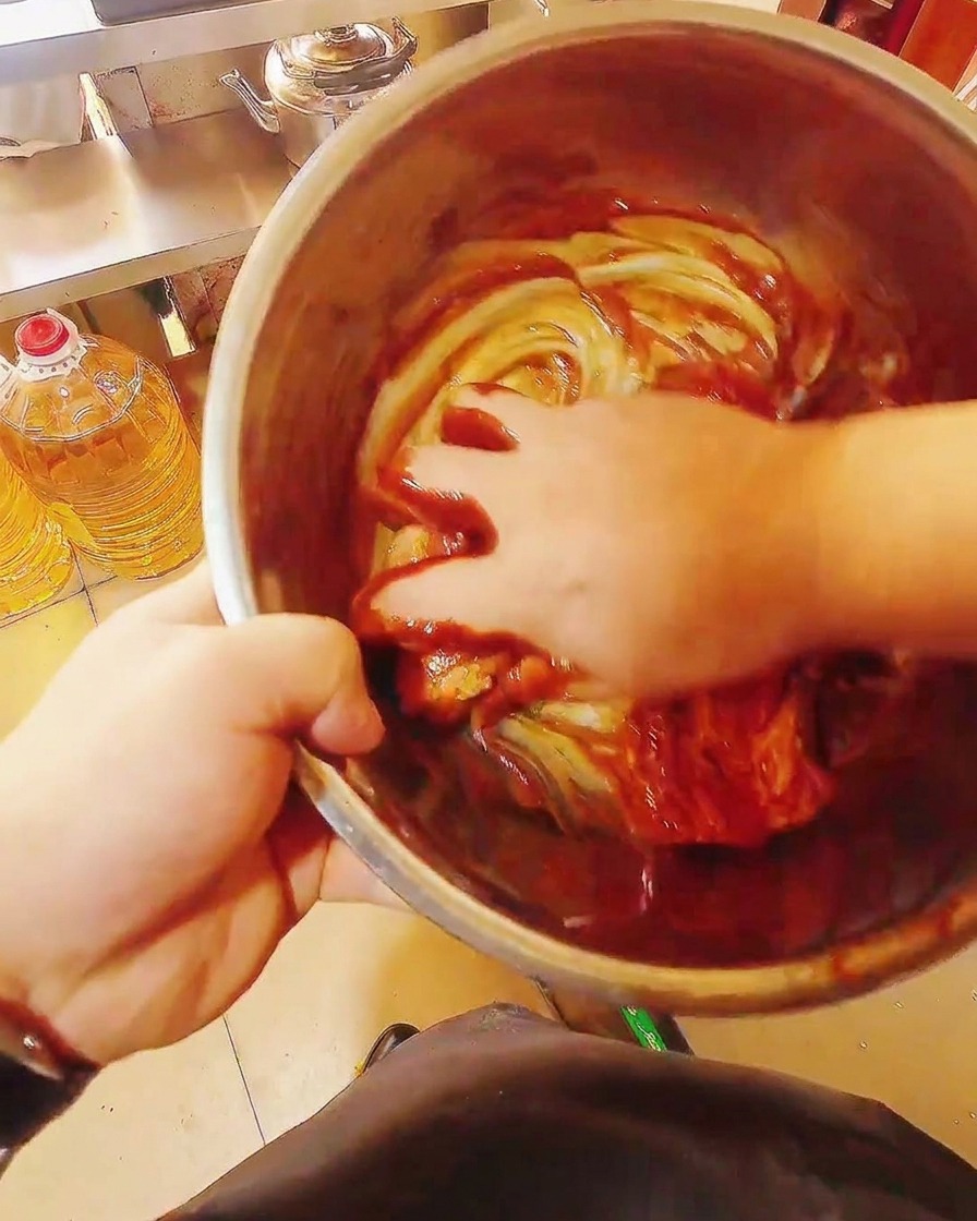 Raw pork belly strips being hand-mixed in a large bowl with a thick, glossy red marinade.