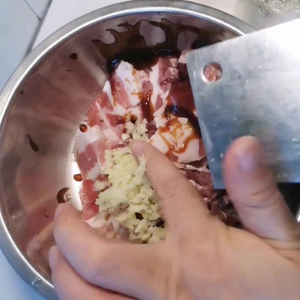 A metal bowl containing sliced pork belly being seasoned with minced garlic and various sauces.