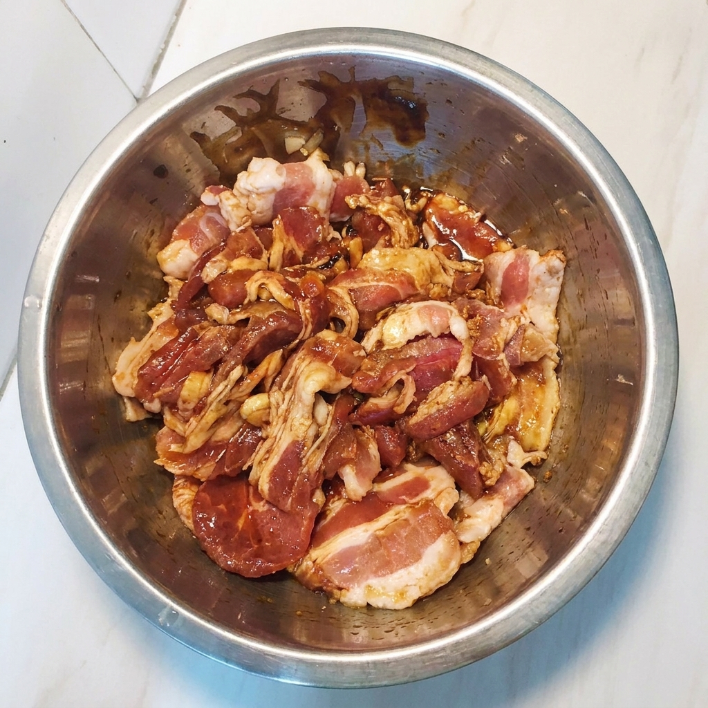 Marinated pork belly slices being tossed and mixed in a metal bowl to ensure even coating.