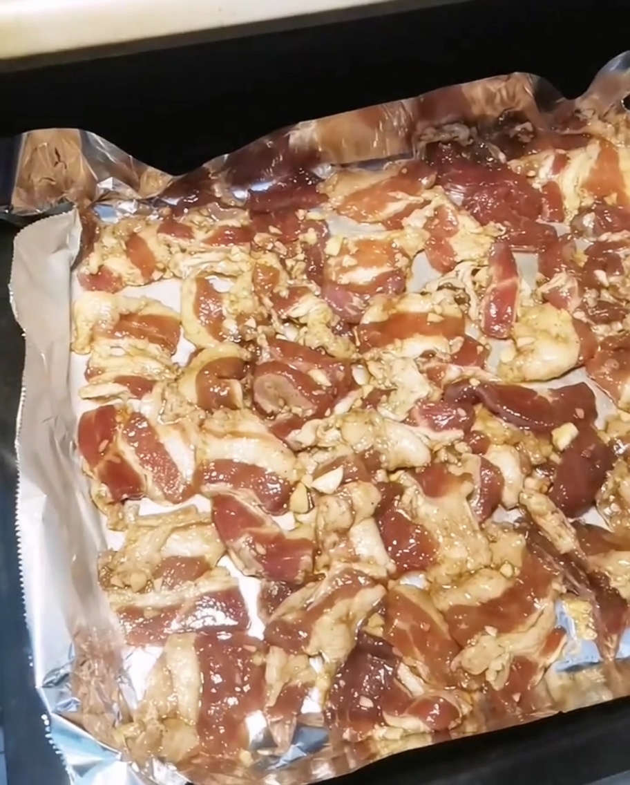 A baking tray filled with seasoned pork belly slices being inserted into a hot oven.