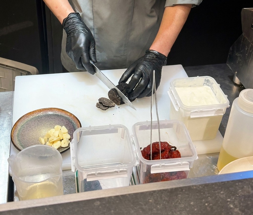 A chef wearing black gloves chopping dried black chilies on a white cutting board.