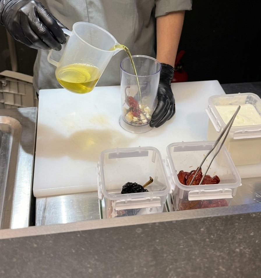 Chef pouring a clear liquid into a tall blender jug containing garlic and chilies.