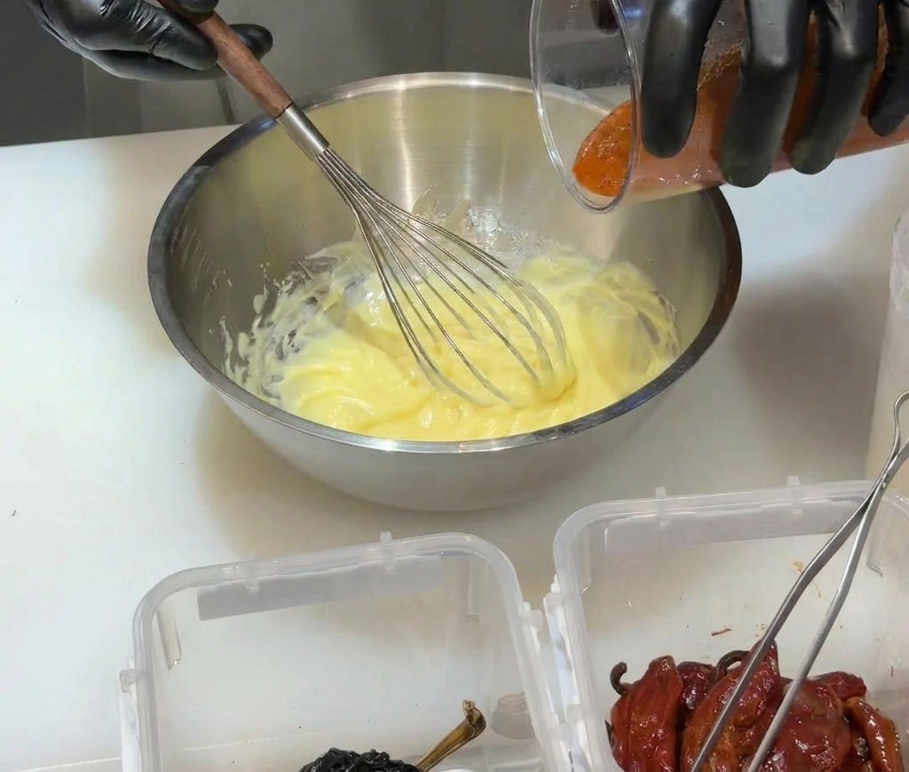 Pouring red blended chili puree into a yellow mayonnaise base in a metal bowl.