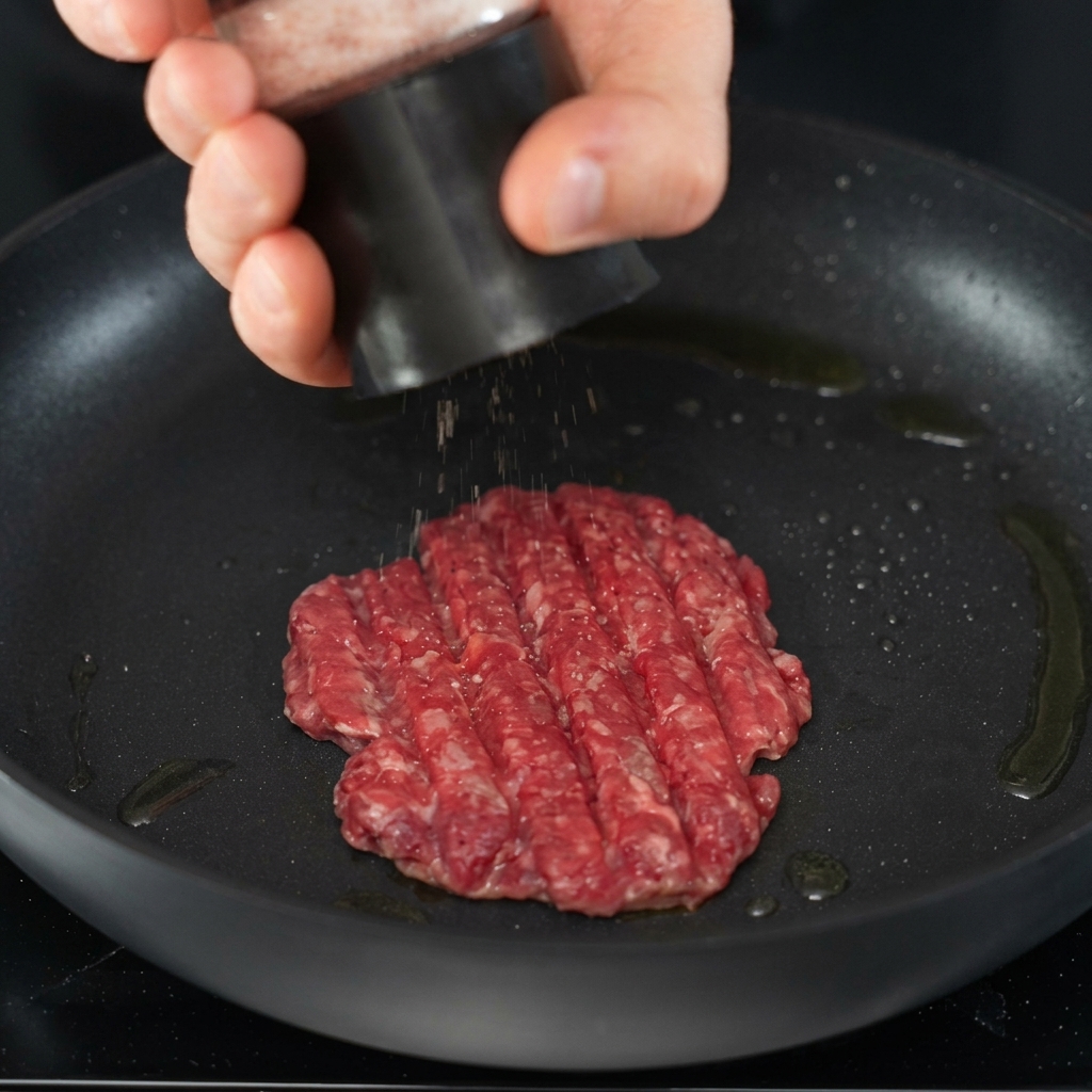 Salt and pepper being ground directly over a searing beef patty in a skillet.
