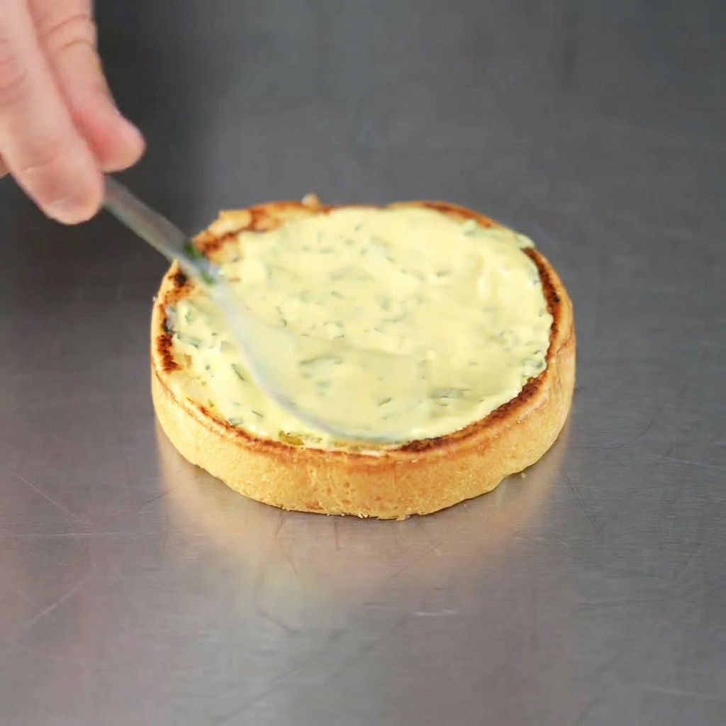 A freshly toasted burger bun being spread with green herb sauce, ready for the patty.