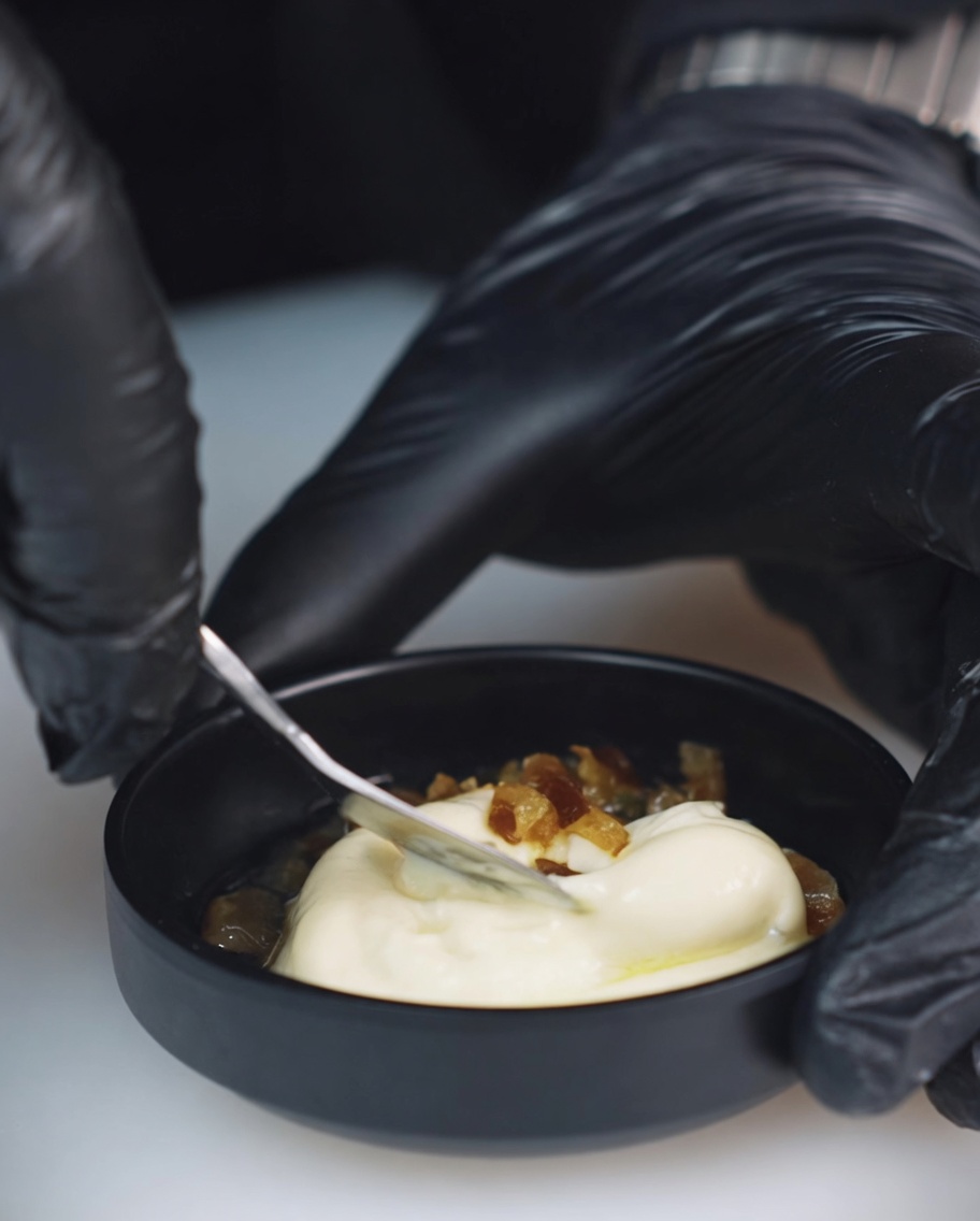 A close-up of a small bowl containing mayonnaise, chopped Mexican chili peppers, and pickle juice being stirred with a metal spoon.