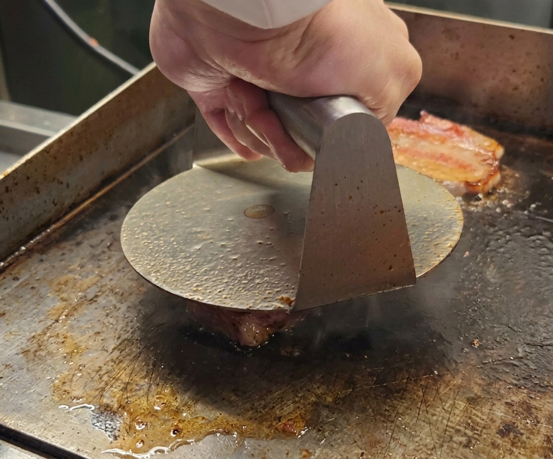A metal burger press flattening a ball of ground Wagyu beef onto a hot cooking surface.