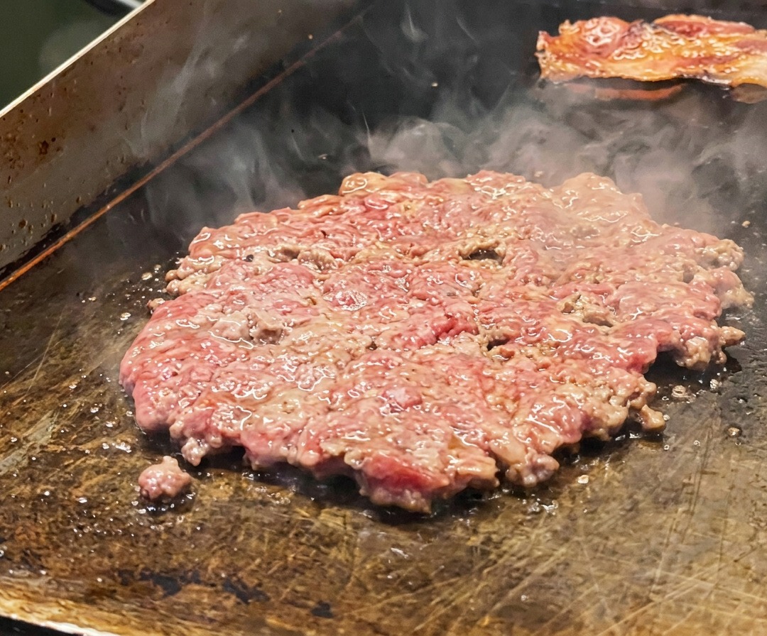 A thin beef patty searing on the griddle, seasoned with salt and topped with onion slices.