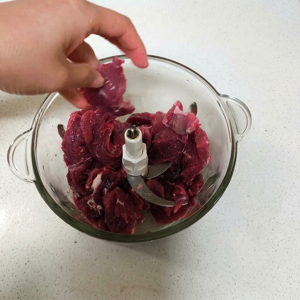 A hand placing pieces of raw red beef into a glass food processor bowl.