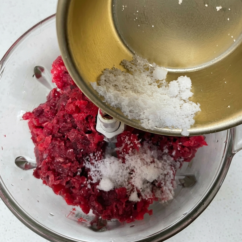 Pouring a white powder mixture of seasonings and starch from a small metal bowl into a food processor with ground beef.