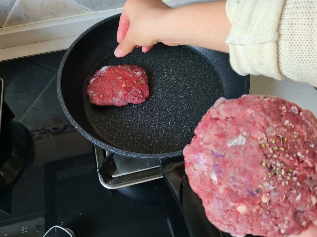 A hand holding a seasoned, raw, circular beef patty while placing another patty into a heated, lightly oiled non-stick frying pan.