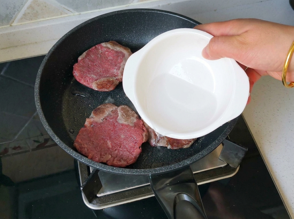 A hand pouring a splash of water from a small white bowl into a frying pan containing three deeply browned beef patties.