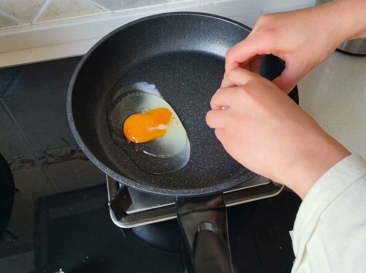 A persons hand cracking a raw egg with a bright yellow yolk into a dark, heated frying pan.