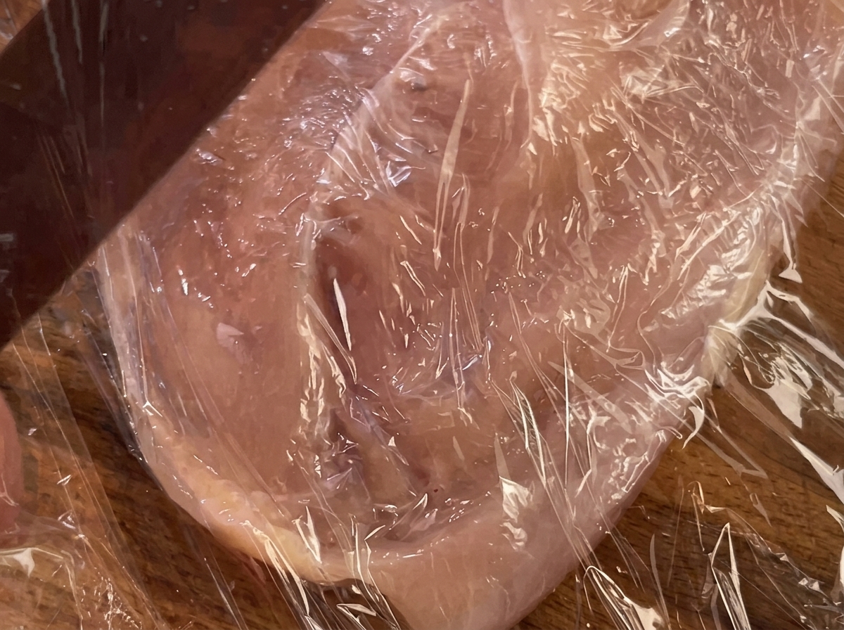 A rolling pin pressing down on a chicken breast covered with plastic wrap.
