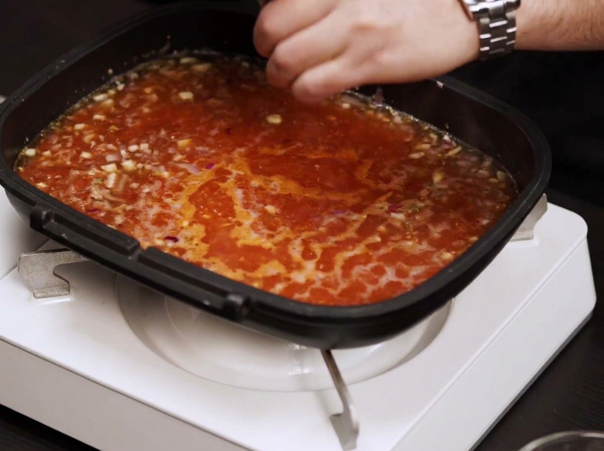 A pan filled with a bubbling red tomato-based sauce while a hand seasons it with salt and pepper.