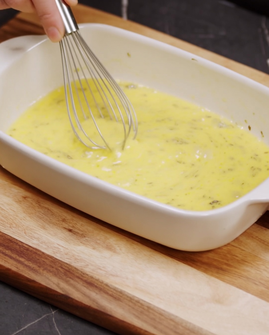 A whisk blending breadcrumbs, grated parmesan cheese, and black pepper in a white ceramic baking dish.