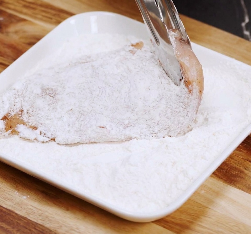 A raw chicken breast fillet being coated in flour on a white rectangular plate using metal tongs.