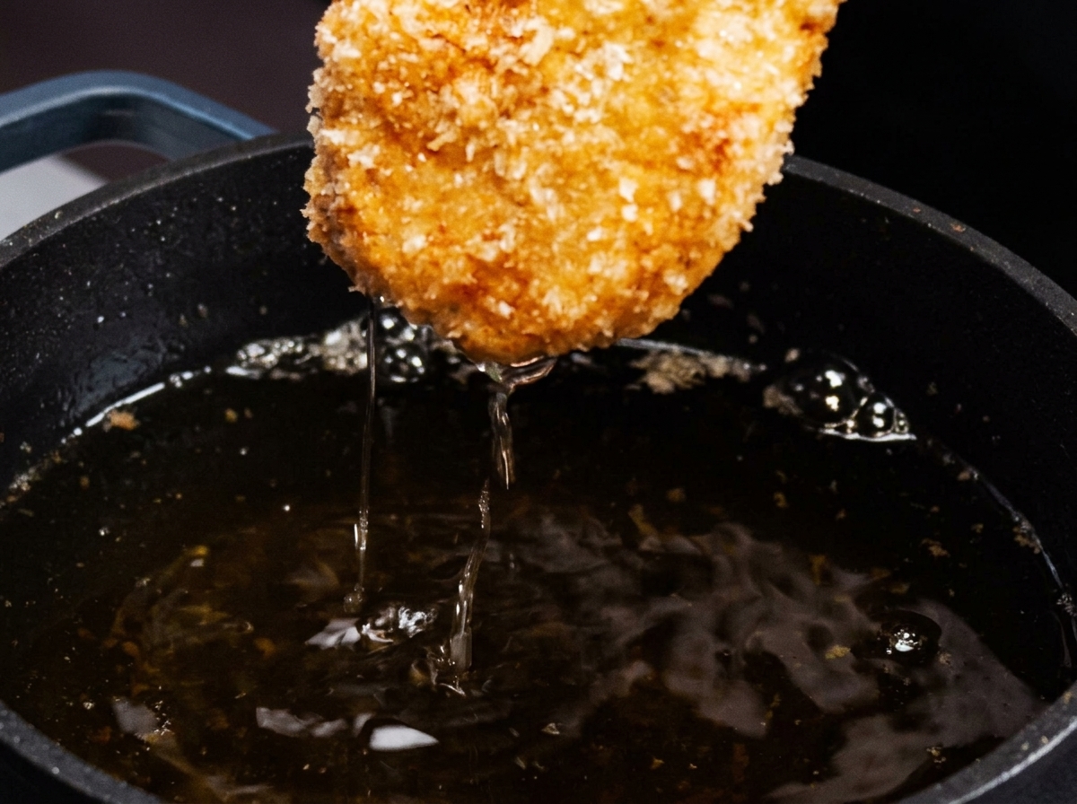 A breaded chicken cutlet being lifted from a pan of hot oil, showing a golden brown crust.