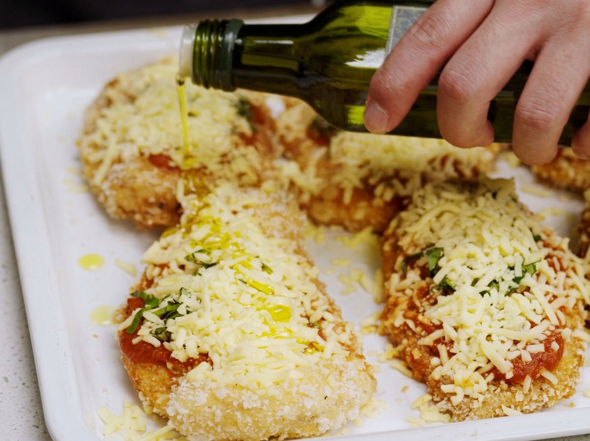 Olive oil being drizzled from a bottle onto assembled Chicken Parmesan on a baking tray.