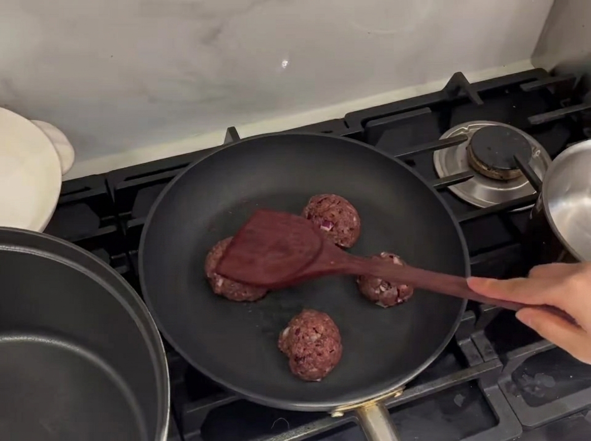 Raw beef meatballs being smashed flat in a hot frying pan with a metal spatula.