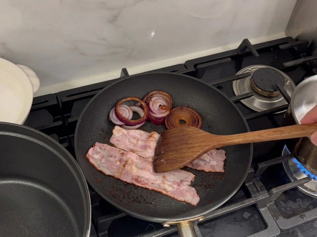 Sizzling bacon strips and red onion rings frying in a pan.