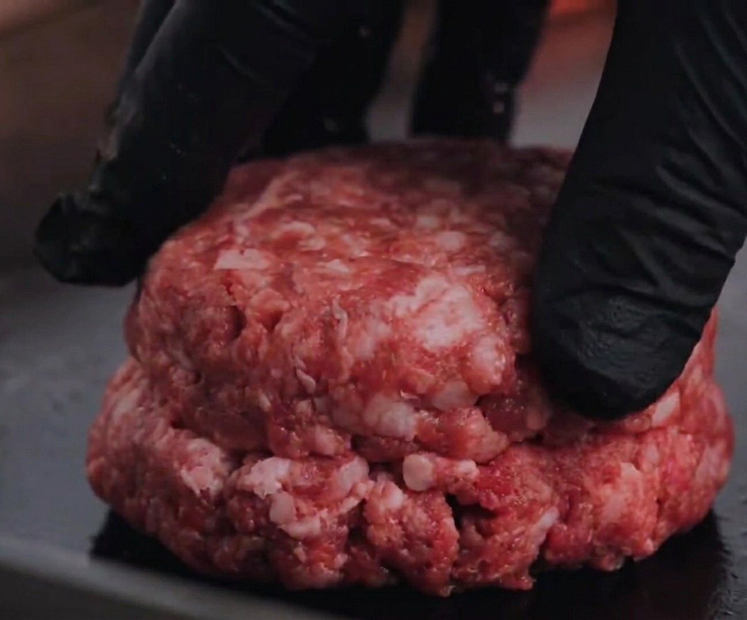 A raw Angus beef patty being pressed onto a hot grill.