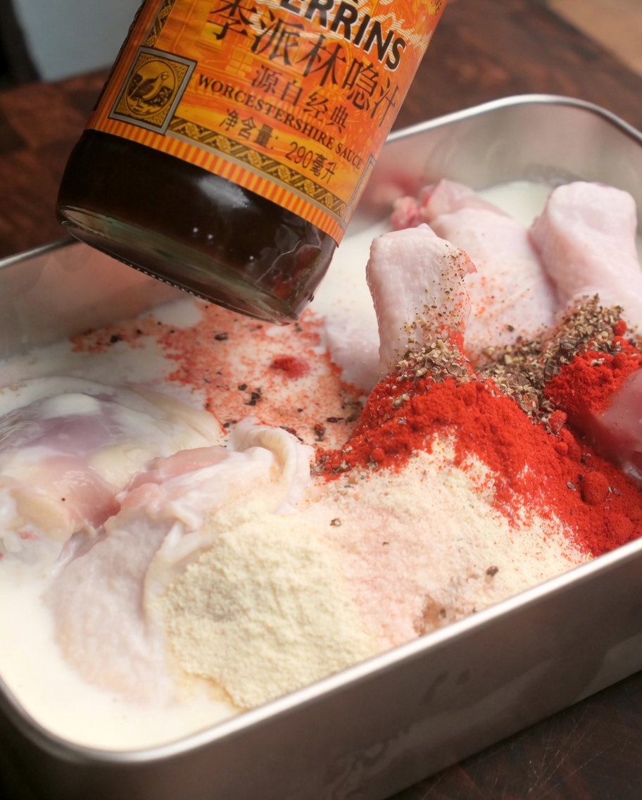 Worcestershire sauce being dripped into a tray of marinated raw chicken drumsticks coated with red spice powders.
