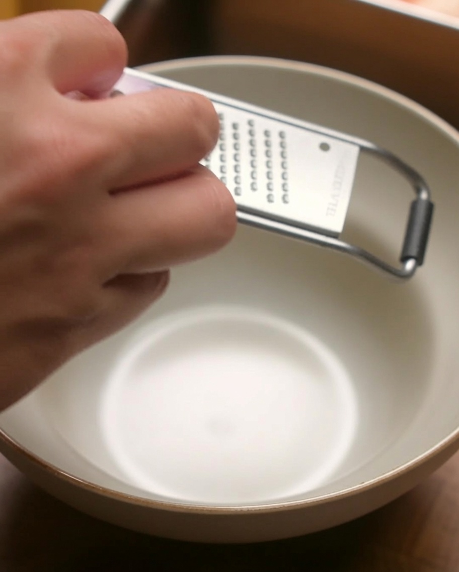 A hand grater positioned over a beige bowl, being used to finely grate a piece of red onion.