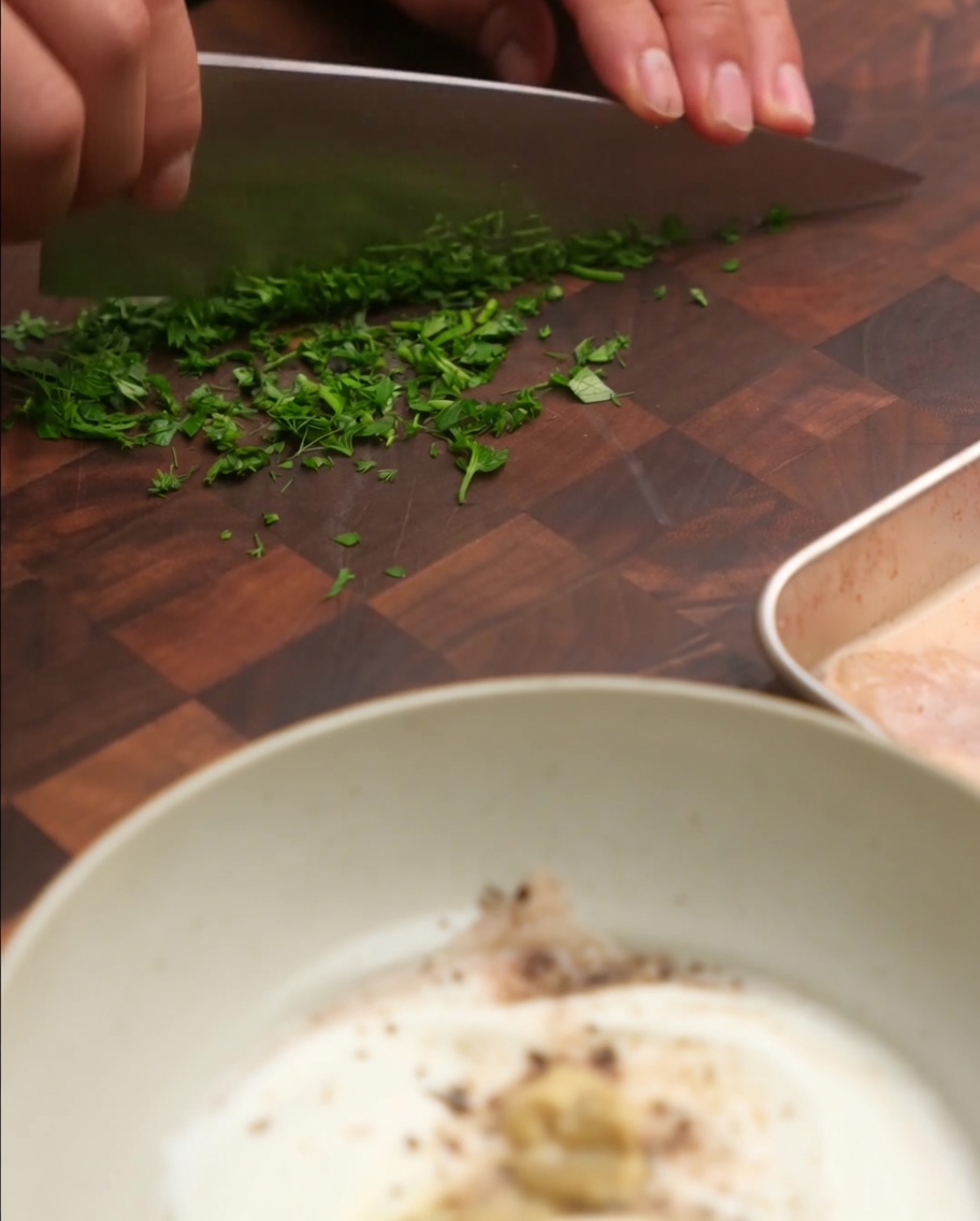 Chopped fresh green chives and parsley being added to a mixing bowl with yogurt and other sauce ingredients.