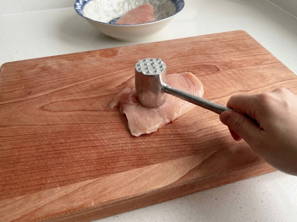A close-up of a meat mallet tenderizing a raw chicken breast on a wooden surface.